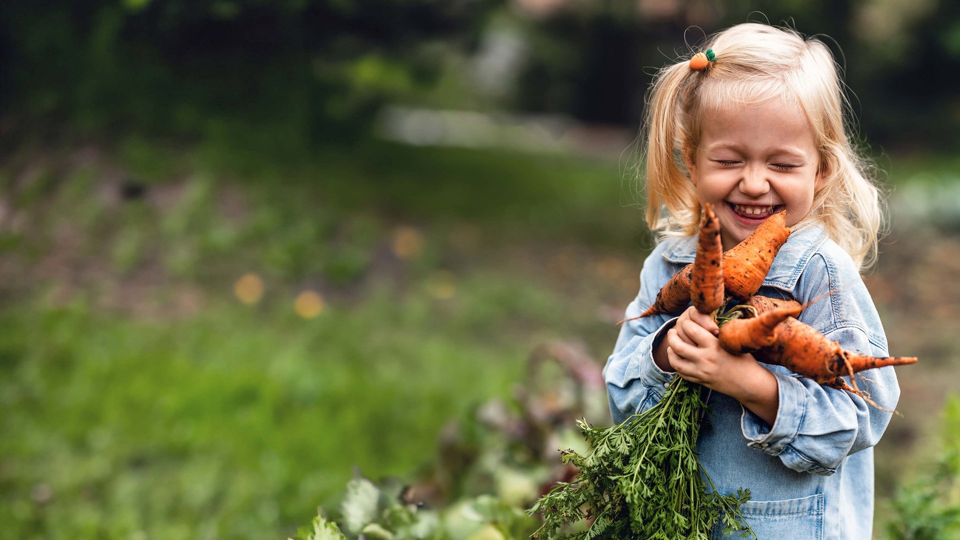 Kinder- und Jugendvorsorge: Ein Mädchen mit blonden Haaren steht im Garten, hält frisch geerntete Karotten in den Händen und lacht.