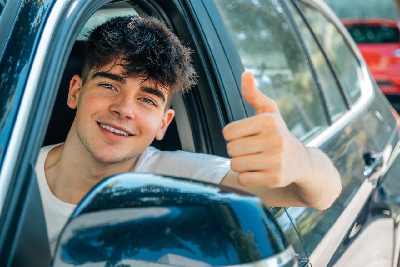 Ein junger Mann, der gerade seinen Führerschein bekommen hat, sitzt auf dem Fahrersitz eines Autos, lächelt in die Kamera und hält einen Daumen hoch.