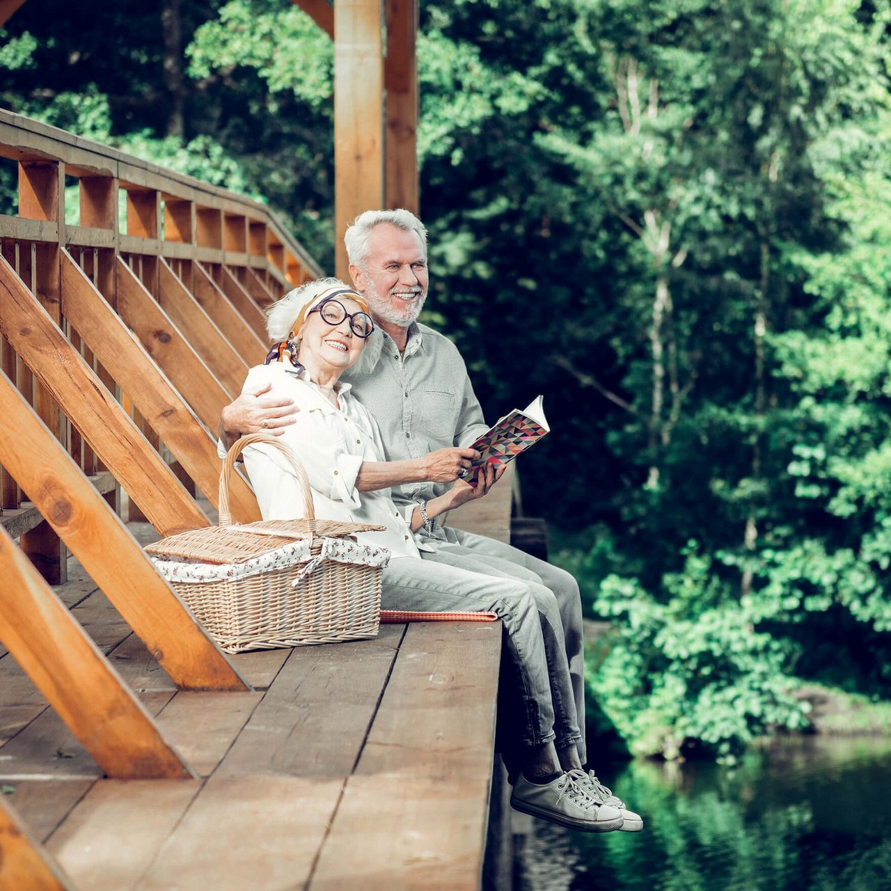 Lachende ältere Frau umarmt lachenden älteren Mann. Sie sitzen gemeinsam auf einer hölzernen Brücke und haben einen Picknickkorb dabei.