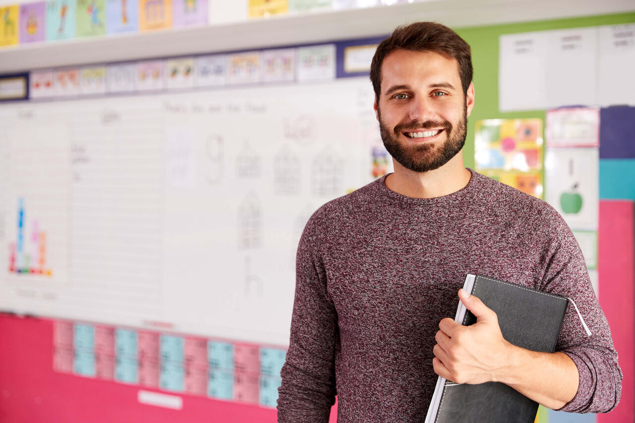 Ein junger Lehrer steht in einer Klasse und lacht in die Kamera. Im Hintergrund ist seine Schulklasse zu sehen. 