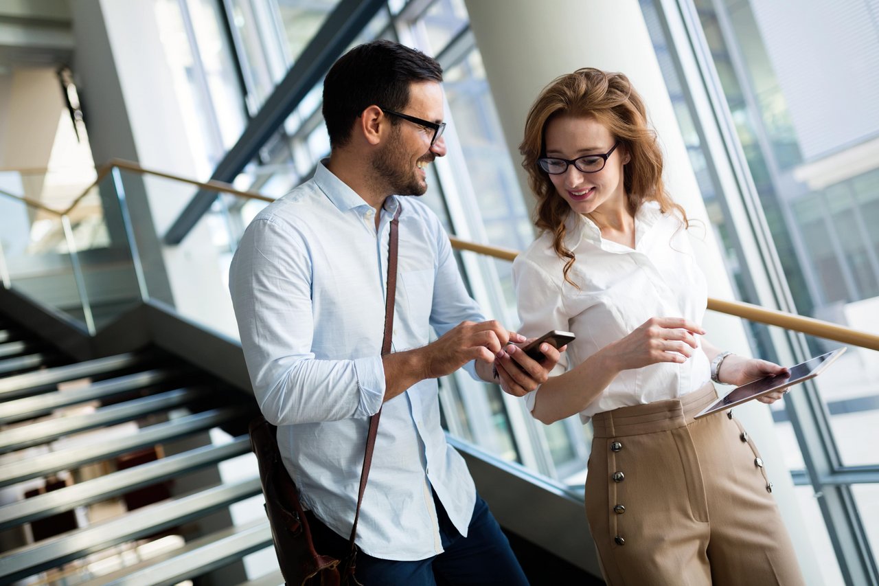 Zwei Kolleg:innen gehen die Stufen im Büro hinunter und unterhalten sich. Der Mann zeigt seiner Kollegin etwas auf seinem Smartphone und lacht dabei.