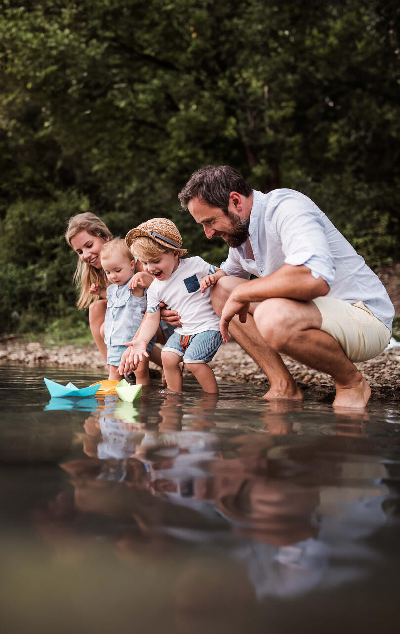 Eine glückliche Familie mit zwei kleinen Kindern steht am Wasser und lässt kleine, gefaltete Boote ins Wasser.