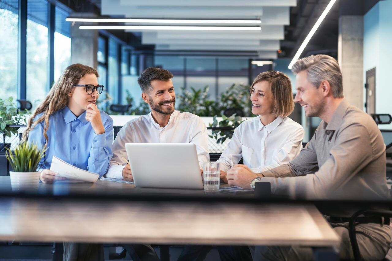 Vier Kolleg:innen sitzen an einem Tisch und halten ein Meeting ab. Eine Frau arbeitet dabei am Laptop.