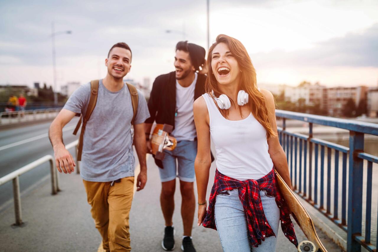 Eine Gruppe junger Leute spaziert lachend mit Rucksäcken und Skateboard in der Hand über eine Brücke.