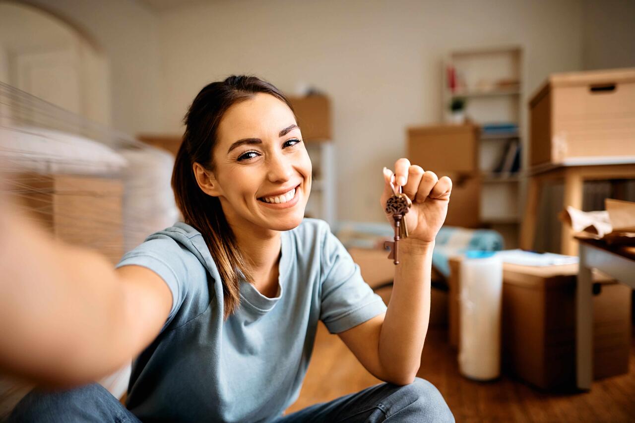 Eine junge Frau macht ein Selfie und lächelt in die Kamera, während sie den Schlüssel ihrer neuen Wohnung in der Hand hält.