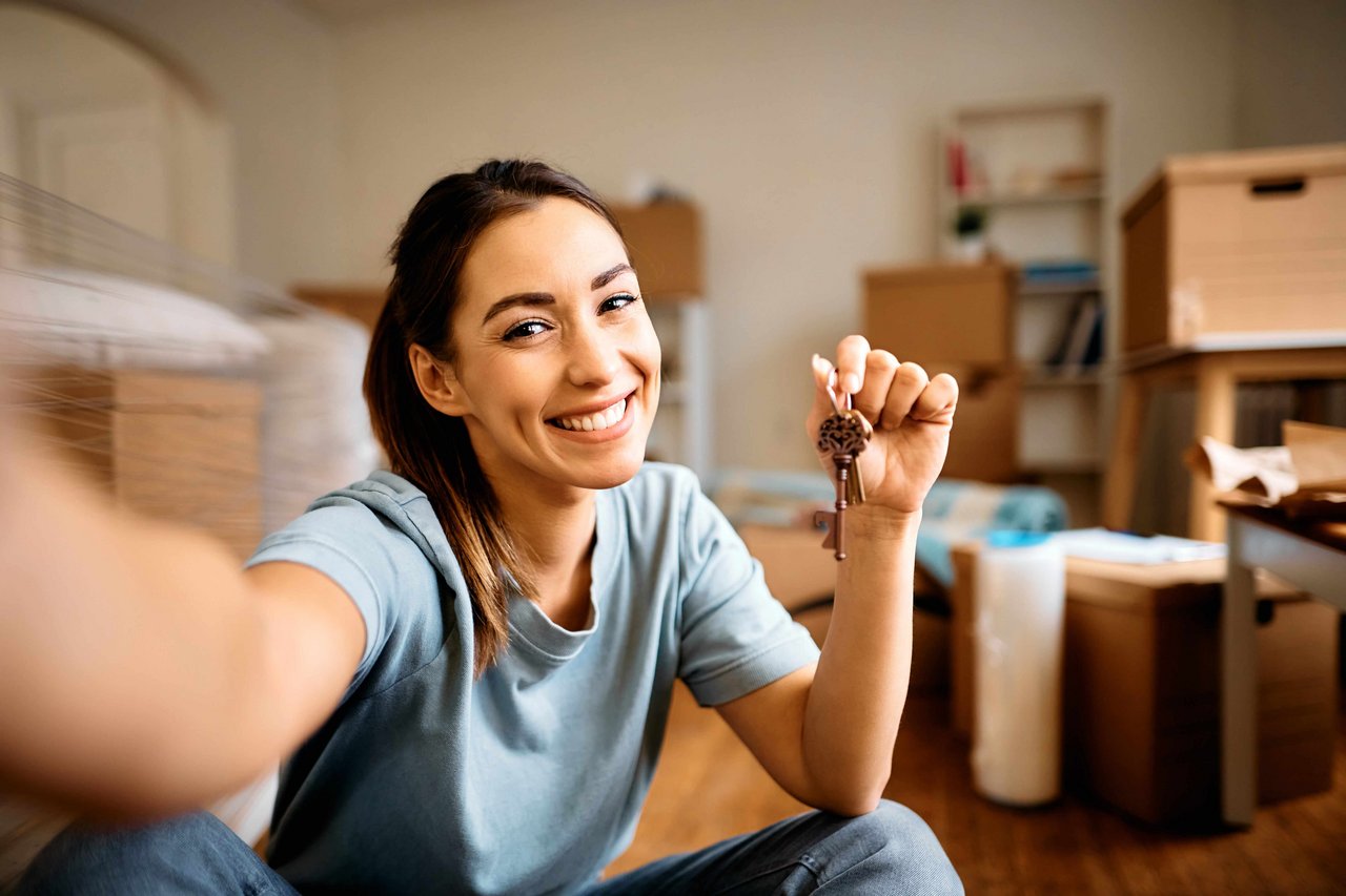 ÖBV Deine Haushaltsversicherung Eine junge Frau macht ein Selfie und lächelt in die Kamera, während sie den Schlüssel ihrer neuen Wohnung in der Hand hält.