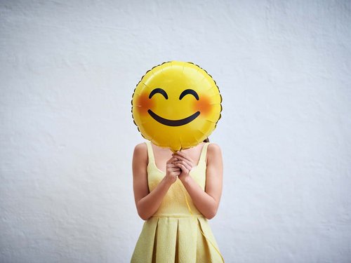 Frau in gelbem Kleid hält sich Smiley-Luftballon vor das Gesicht.