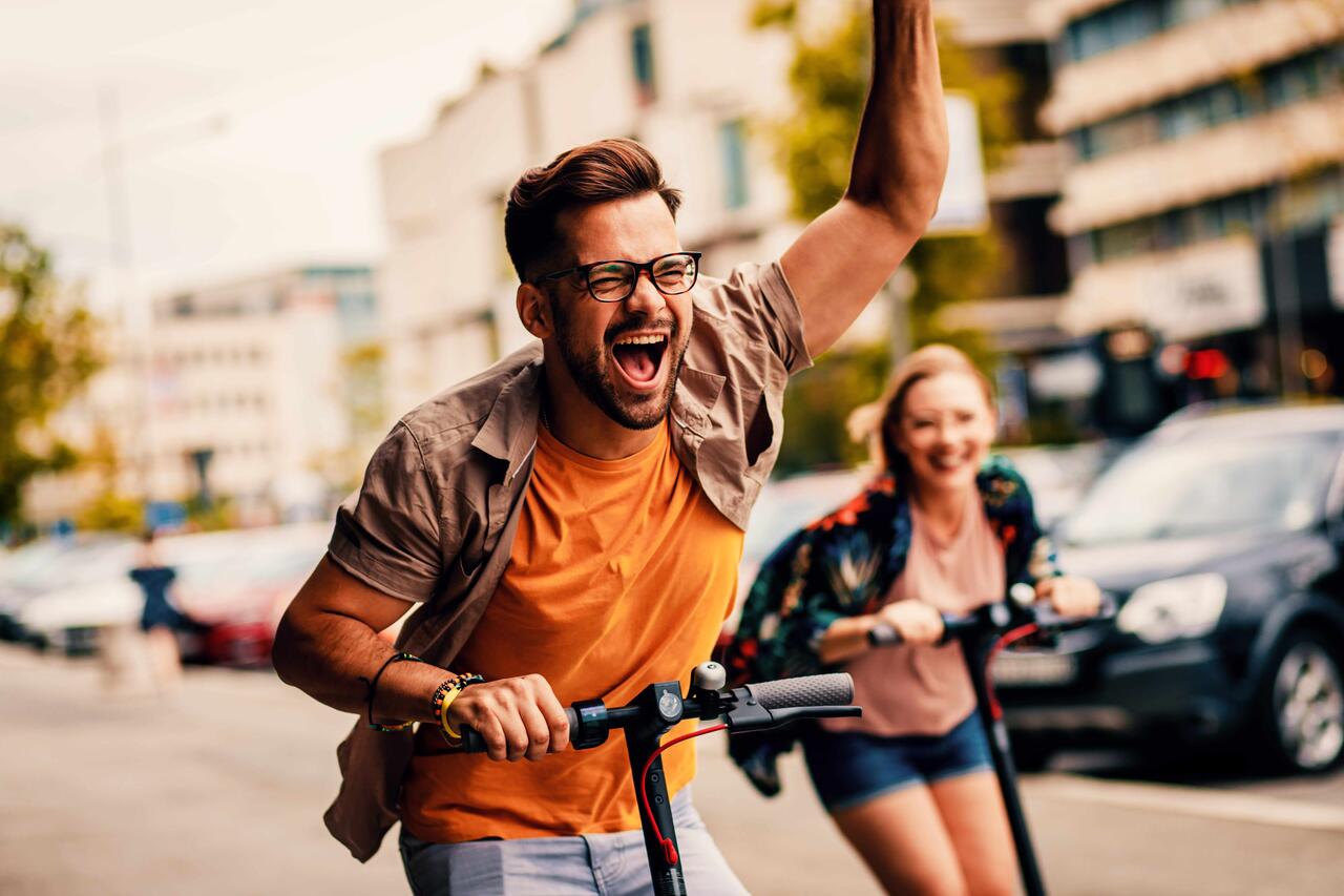Zwei junge Erwachsene fahren auf E-Scootern die Straße entlang.