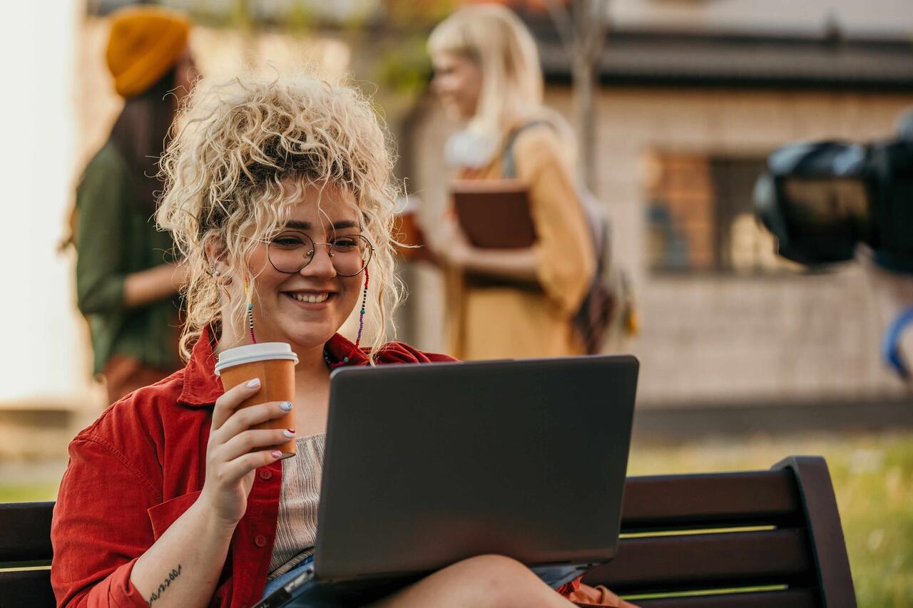 Eine junge Frau sitzt mit ihrem Laptop und einem Kaffee in der Hand auf einer Parkbank und kümmert sich um Bewerbungen.