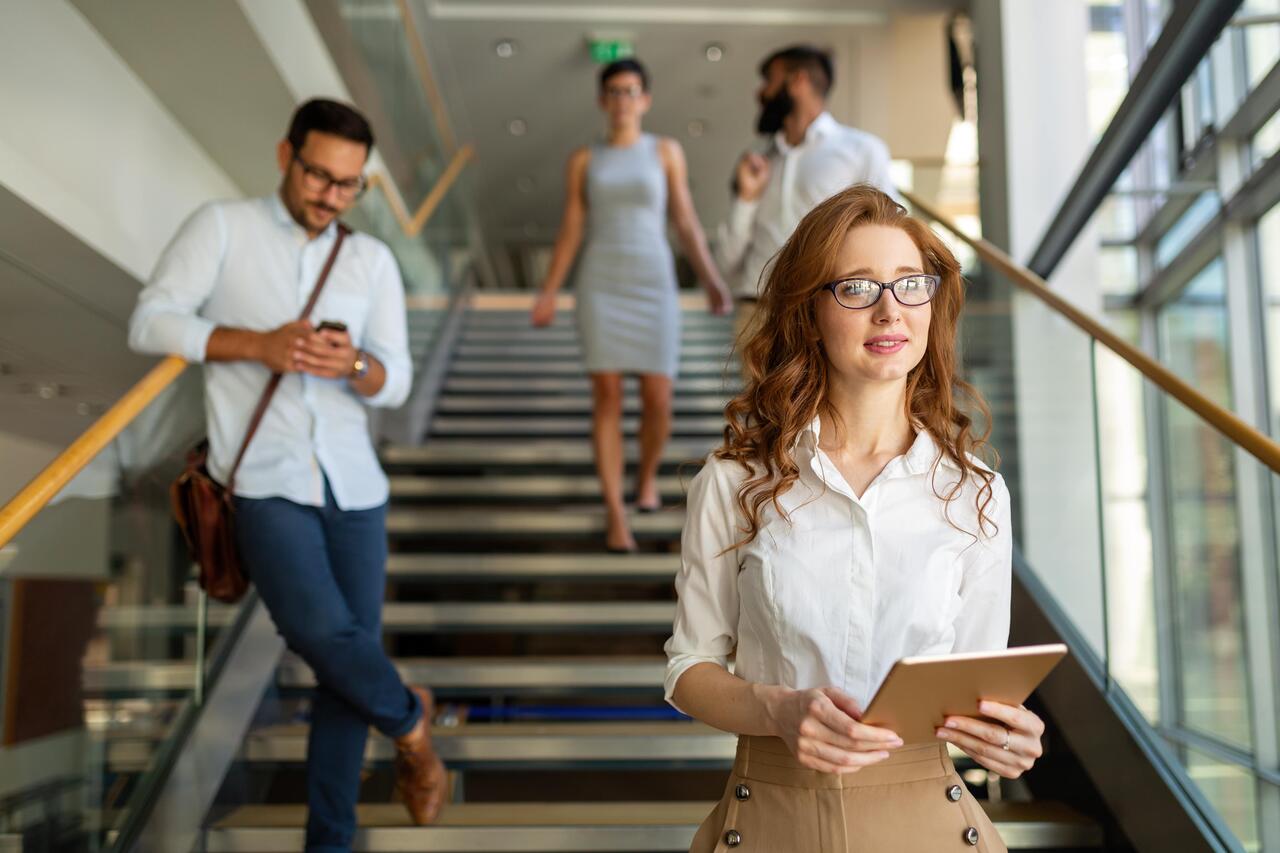 Eine junge Mitarbeiterin ist gerade die Stufen im Büro runtergegangen und lächelt. Im Hintergrund sind drei weitere Arbeitskolleg:innen auf der Stiege zu sehen.