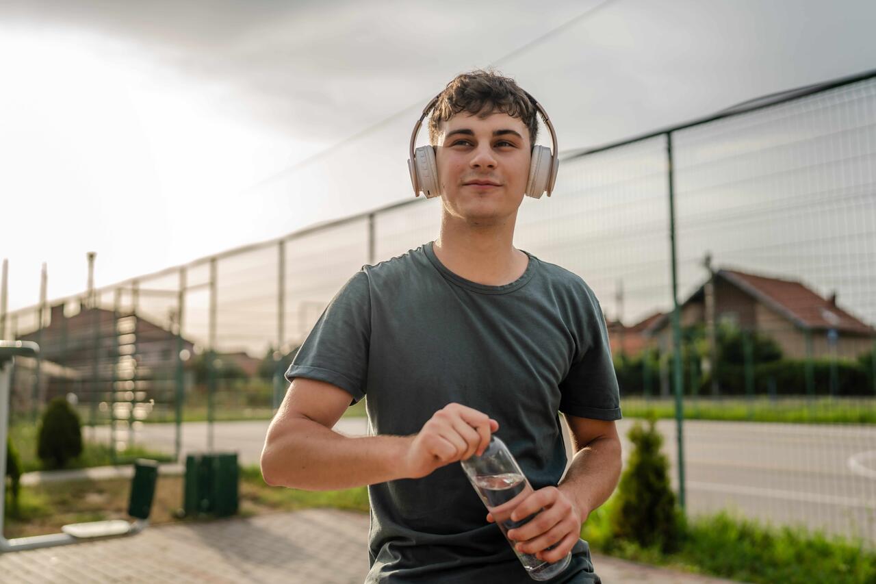 Ein junger Mann macht von seinem Outdoor-Training Pause und hält eine Wasserflasche in der Hand. Er hört dabei Musik.