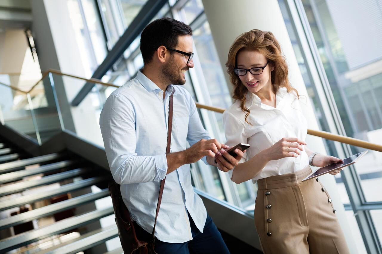 Zwei Kolleg:innen gehen die Stufen im Büro hinunter und unterhalten sich. Der Mann zeigt seiner Kollegin etwas auf seinem Smartphone und lacht dabei.