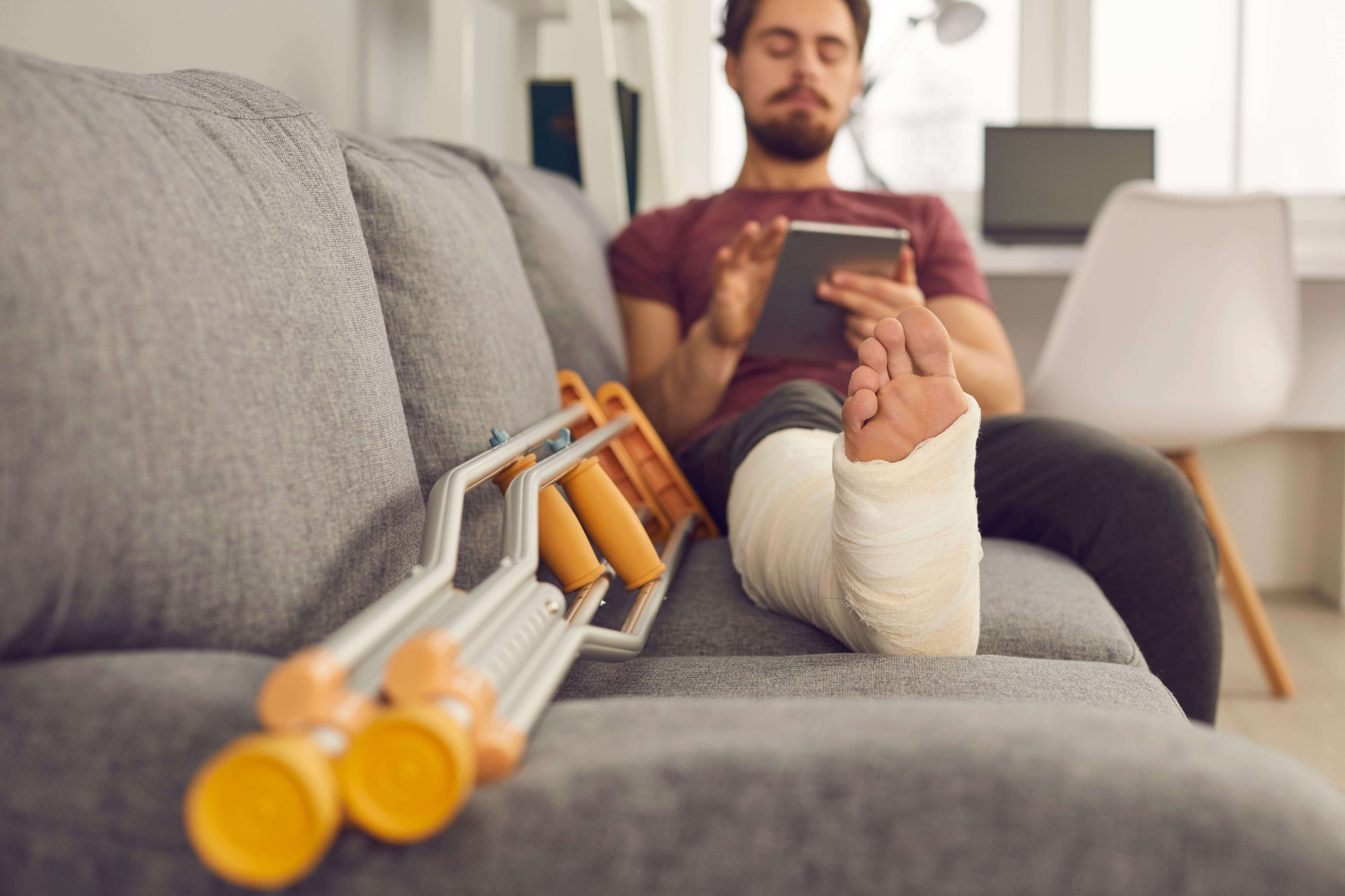 Ein Mann liegt mit einem Gips-Bein auf der Couch. Daneben liegen zwei Krücken.