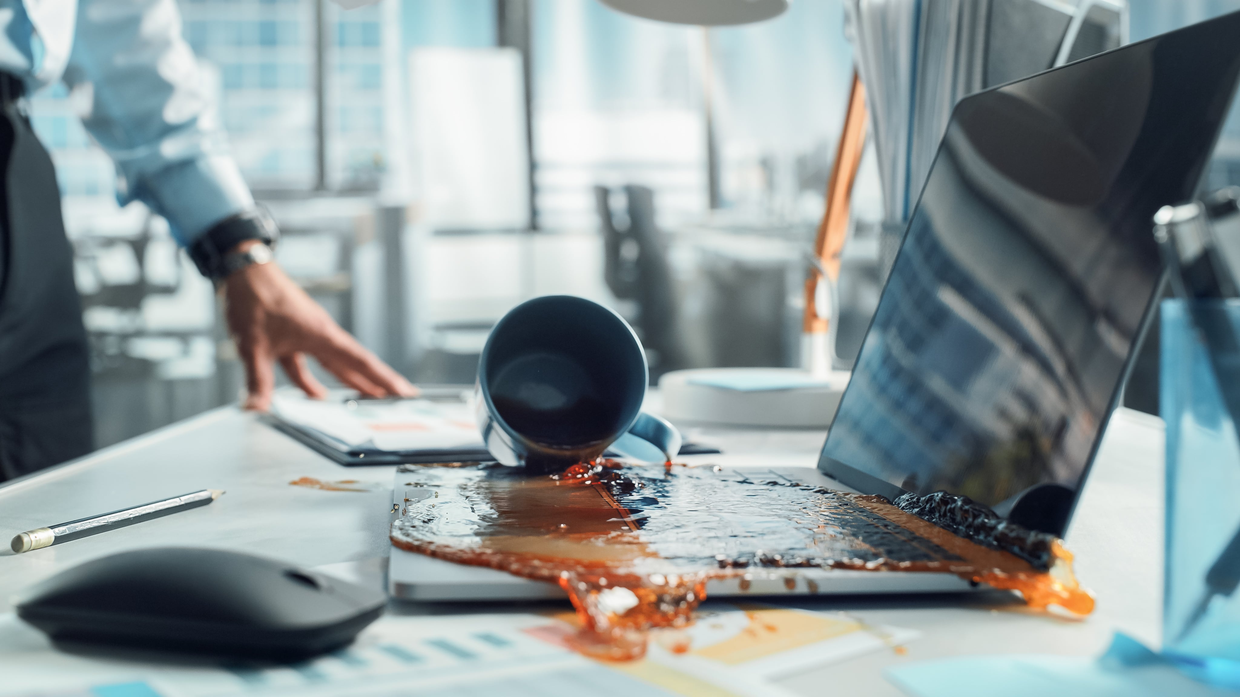 Ein Laptop steht auf einem Schreibtisch. Eine umgekippter Kaffeebecher liegt daneben und der Kaffee wurde über den Laptop verschüttet.