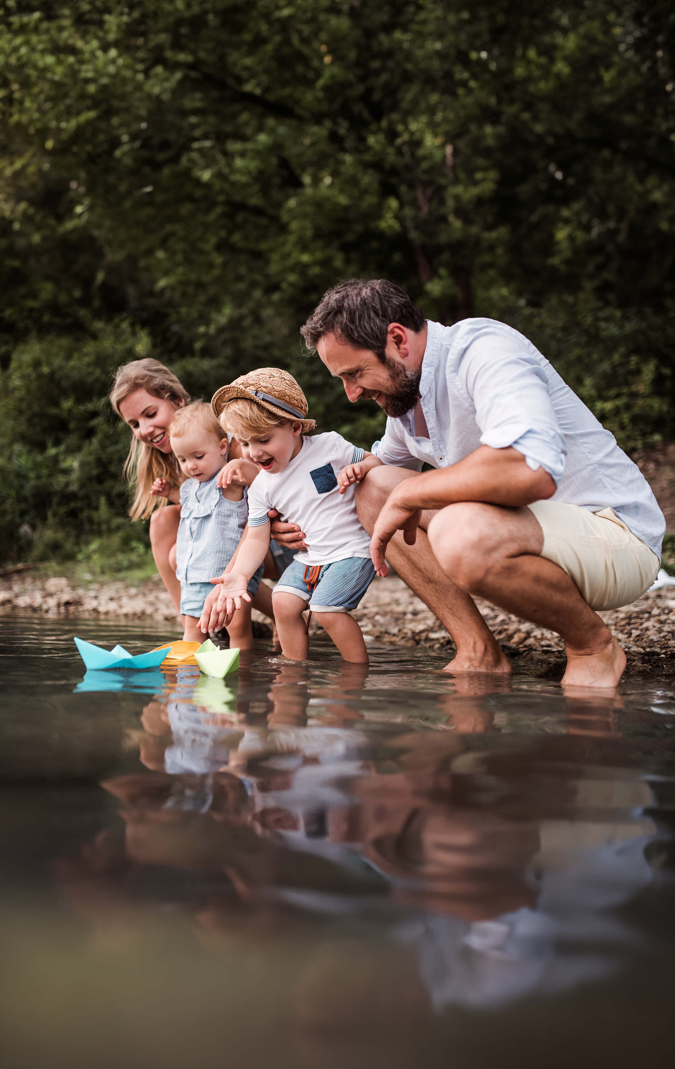 Eine glückliche Familie mit zwei kleinen Kindern steht am Wasser und lässt kleine, gefaltete Boote ins Wasser.