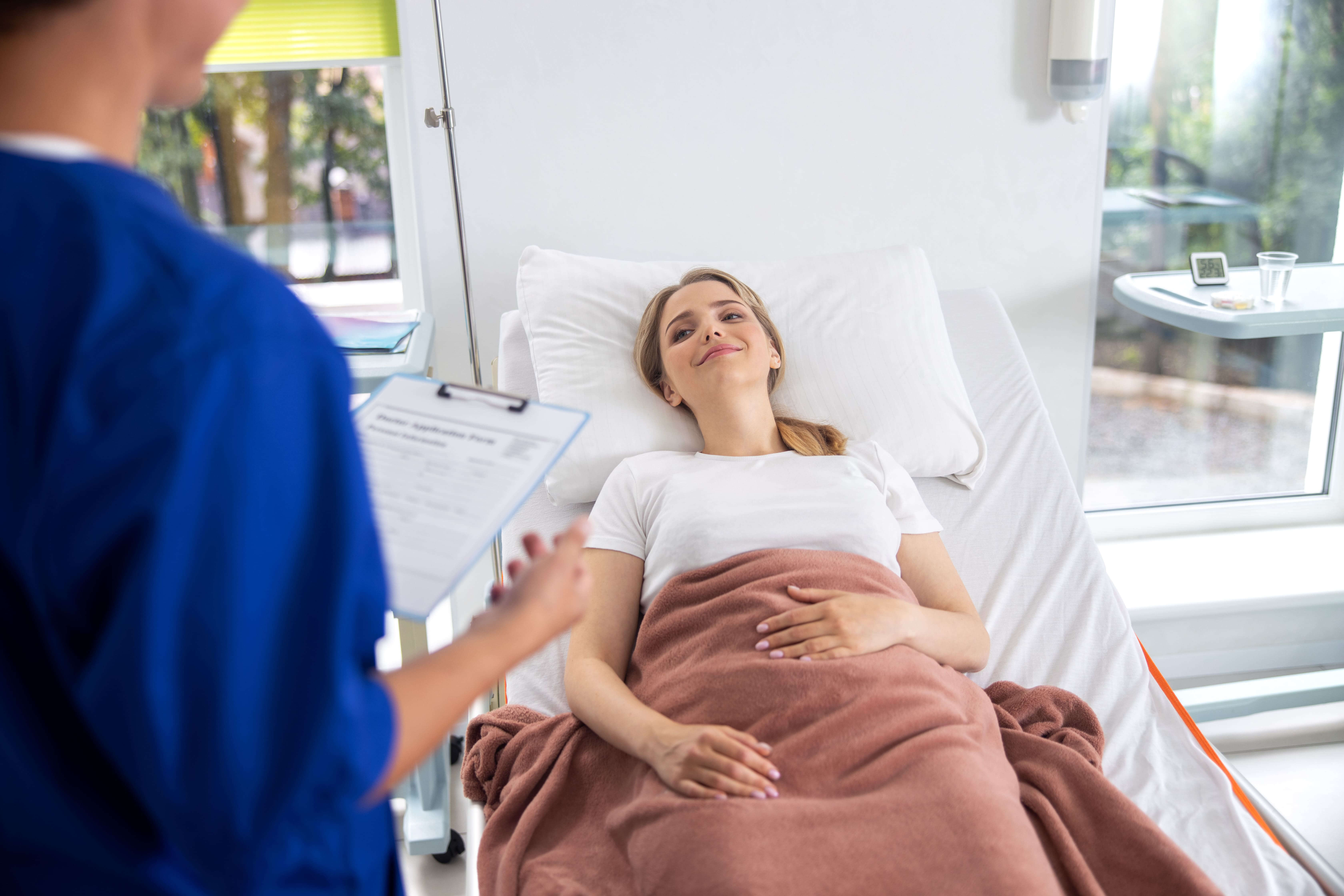 Eine junge Frau liegt in einem Krankenbett und lächelt. Eine Ärztin oder ein Arzt spricht mit ihr.