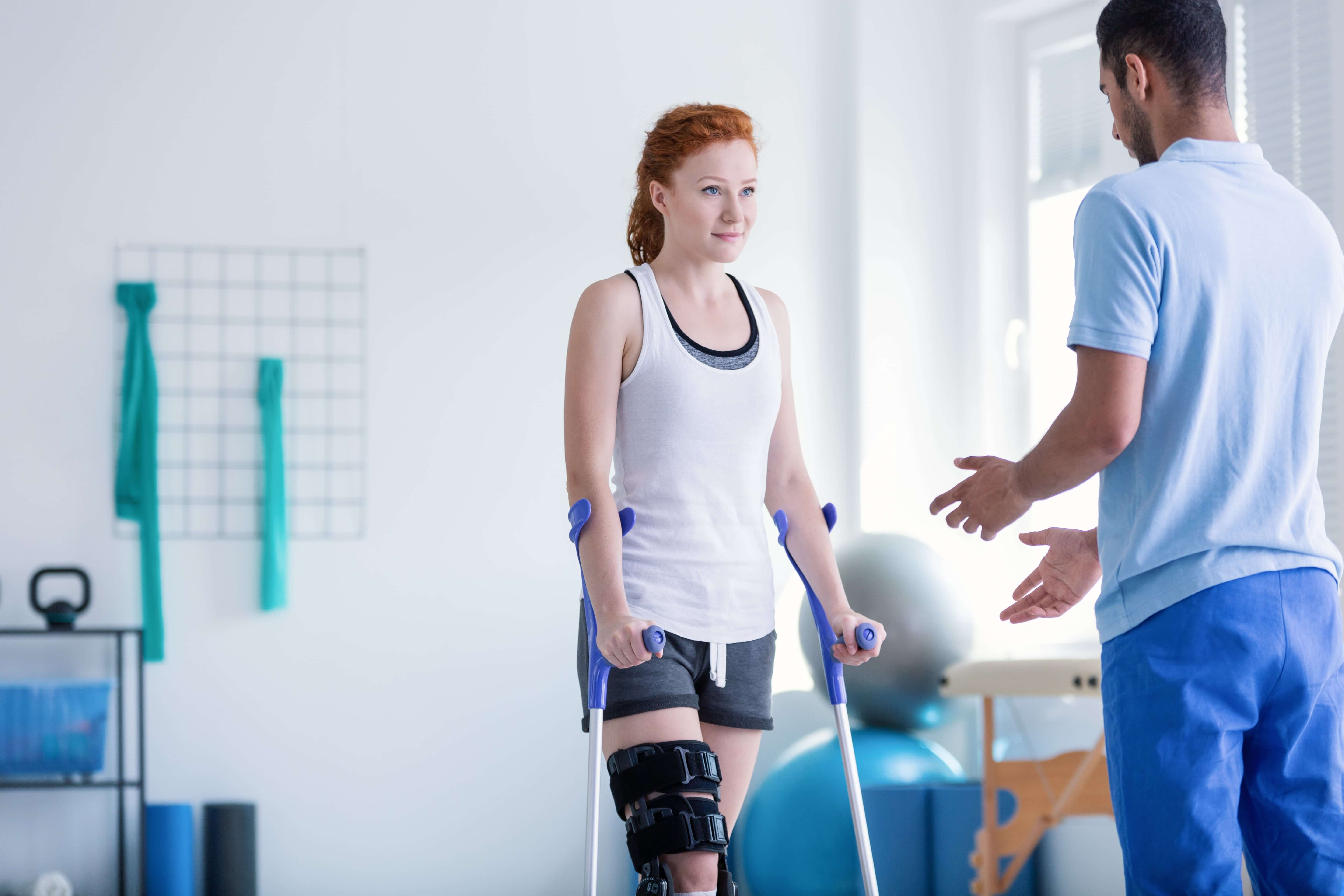 Eine junge Frau steht mit Krücken und einer Schiene am Bein in einem Behandlungsraum. Ein Physiotherapeut steht vor ihr und gibt Instruktionen.