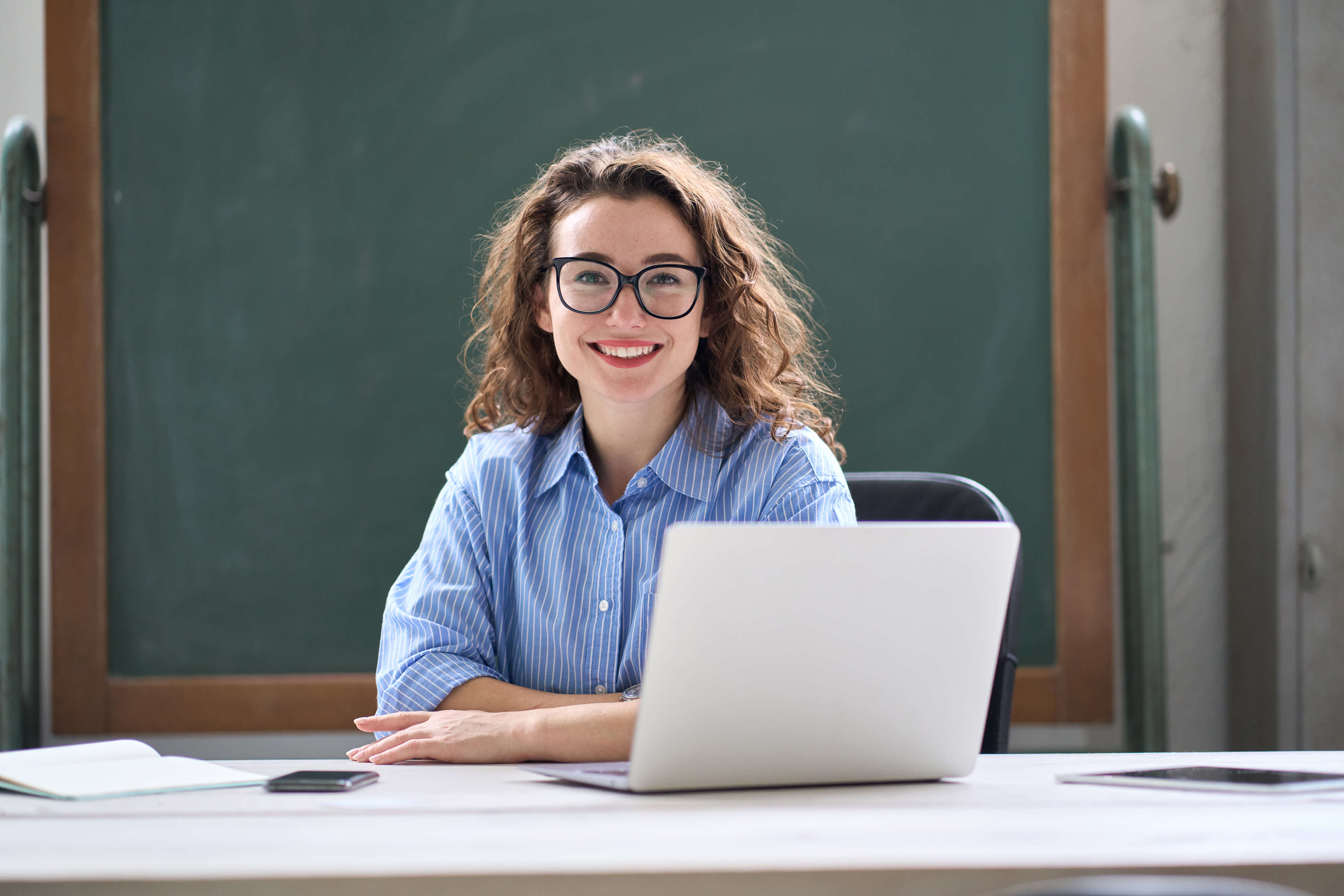 Eine junge Lehrerin und ein junger Lehrer stehen im Klassenzimmer mit dem Rücken zur Tafel und lächeln.